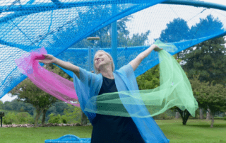 A woman in a blue cardigan dances with pink and green chiffon ribbons outdoors